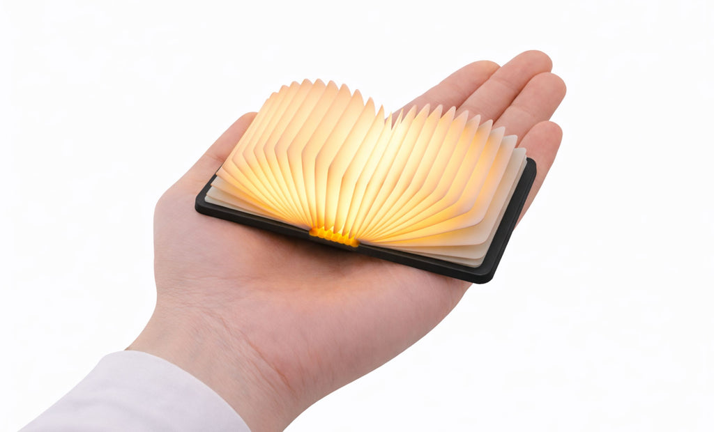Gingko Palm Edition Smart Book Light in graphite/black open and glowing warm amber light in the palm of a hand — shows scale and glow