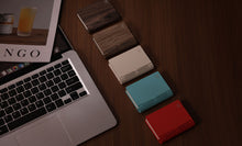 Load image into Gallery viewer, Four Gingko Palm Edition book lights closed and laid flat on a wooden desk beside a laptop — walnut, cream, teal and red 
