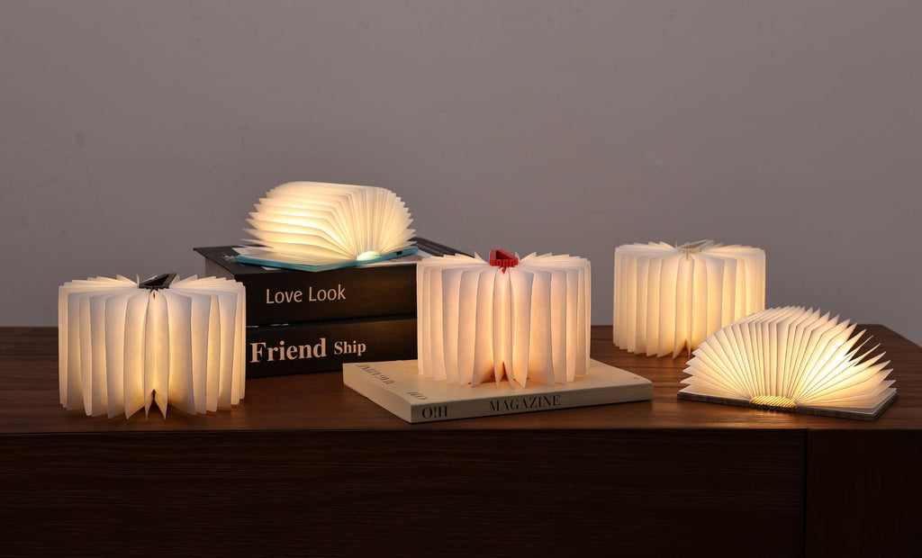 Multiple Gingko Smart Book Lights of various sizes open and glowing on a wooden surface with books in moody ambient lighting