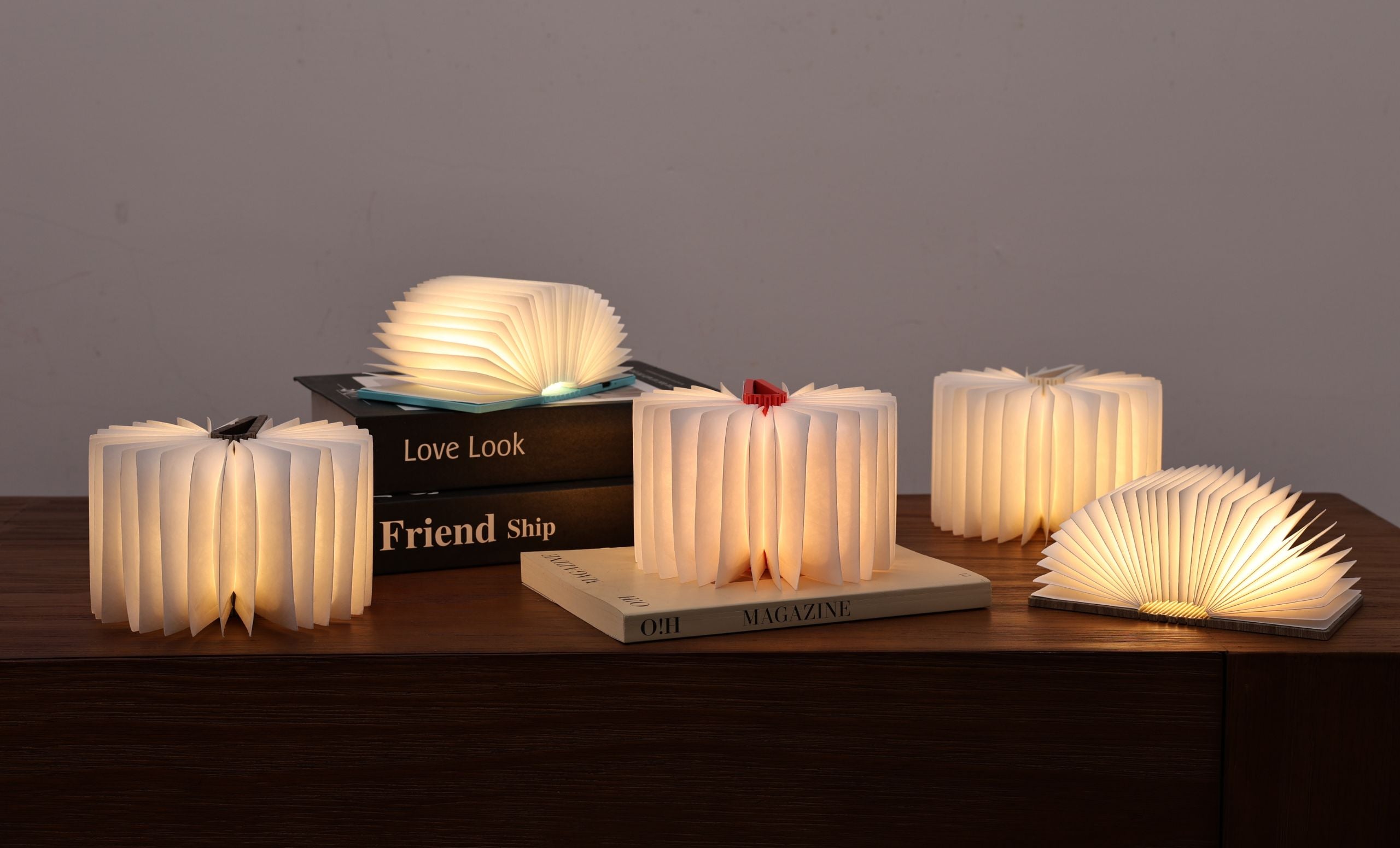 Multiple Gingko Smart Book Lights of various sizes open and glowing on a wooden surface with books in moody ambient lighting