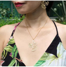 Load image into Gallery viewer, Model wearing RAS Ginkgo Biloba gold pendant and ginkgo leaf drop earrings outdoors with floral dress
