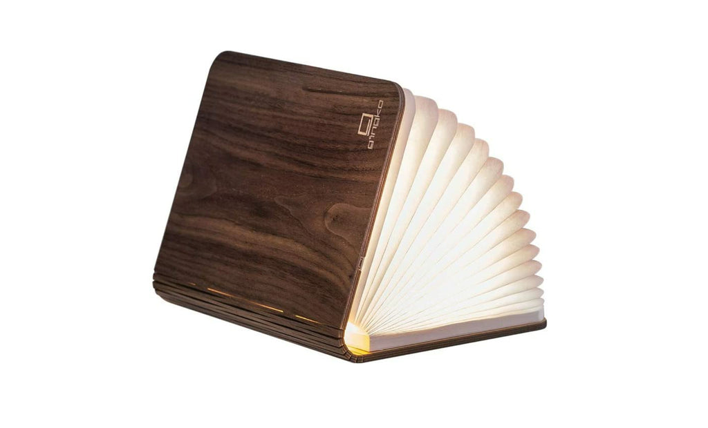 Gingko Smart Book Light in walnut, fully open at 180 degrees showing warm ambient light on desk  book light made of wood, and Tyvek paper, opened to illuminate a warm soft light, placed on a desk next to a notebook and a pen.